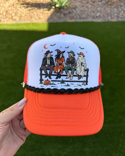 A hand holds an orange and white trucker hat with a black braided trim and a white front panel featuring a cartoon illustration of three Halloween characters sitting on a bench.