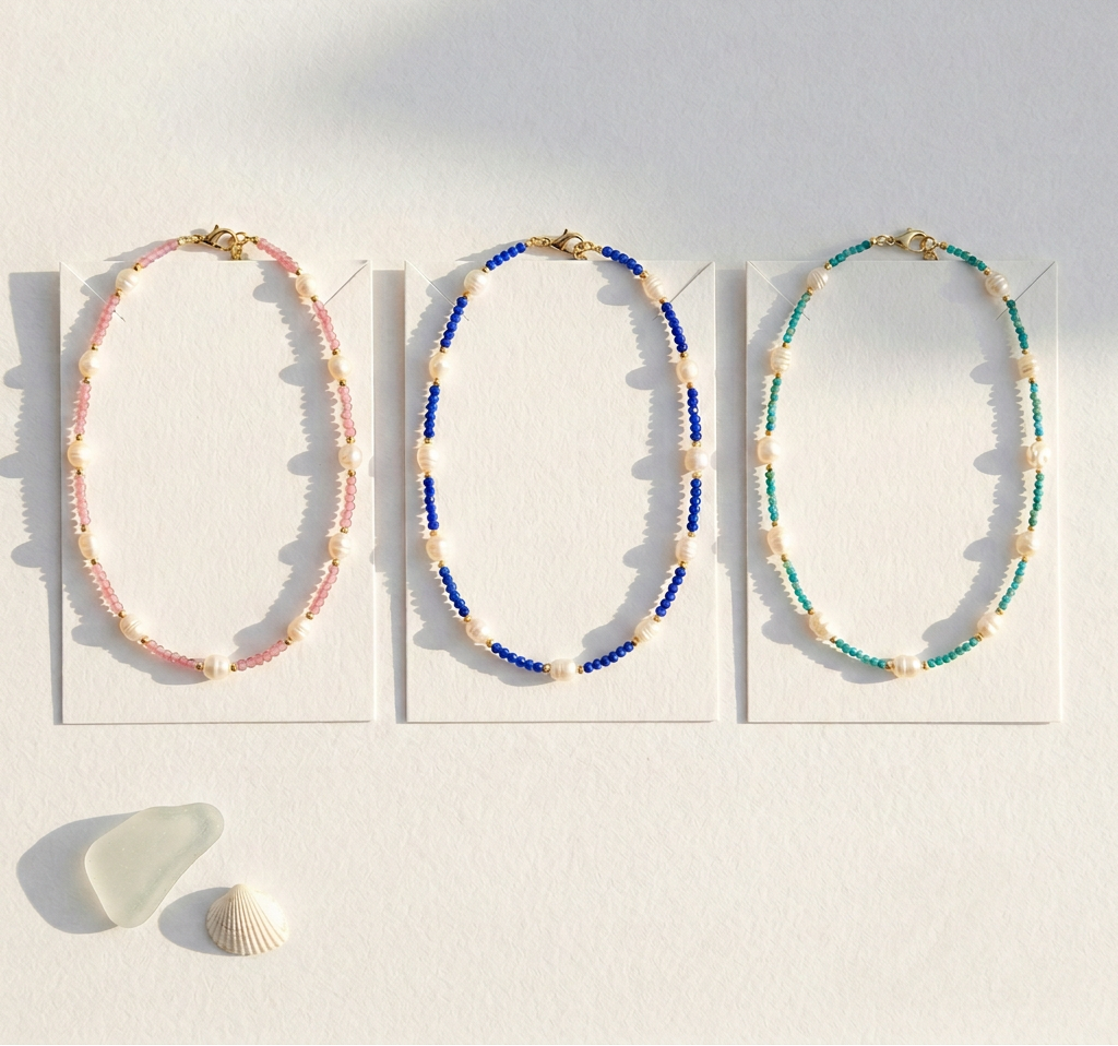 Three beaded necklaces on white cards with a shell on a sandy background