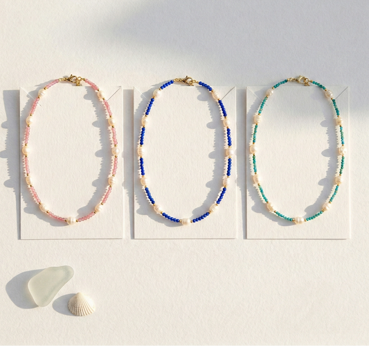 Three beaded necklaces on white cards with a shell on a sandy background