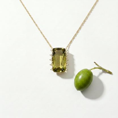 Gold necklace with a green gemstone pendant next to a real green olive on a white background
