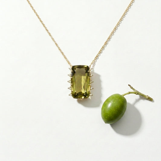 Gold necklace with a green gemstone pendant next to a real green olive on a white background