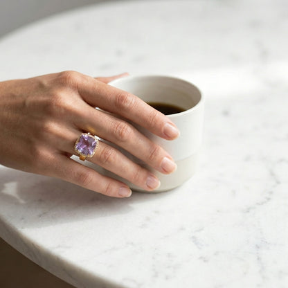 Hand wearing a ring with a purple gemstone holding a white mug on a light surface
