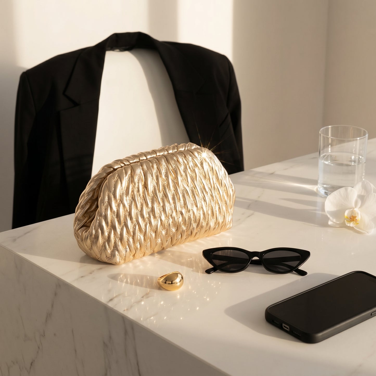 Gold clutch bag on a marble surface with sunglasses, phone, and glass in the background